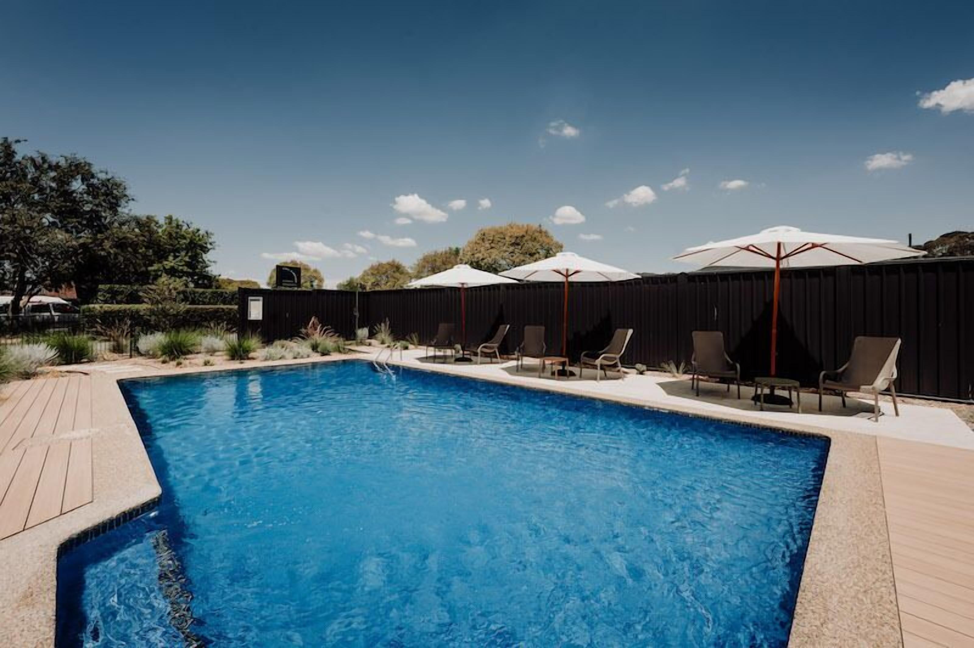 Una piscina rectangular con agua azul cristalina, rodeada por una terraza de madera. Hay sillas de descanso y sombrillas a lo largo de la piscina.