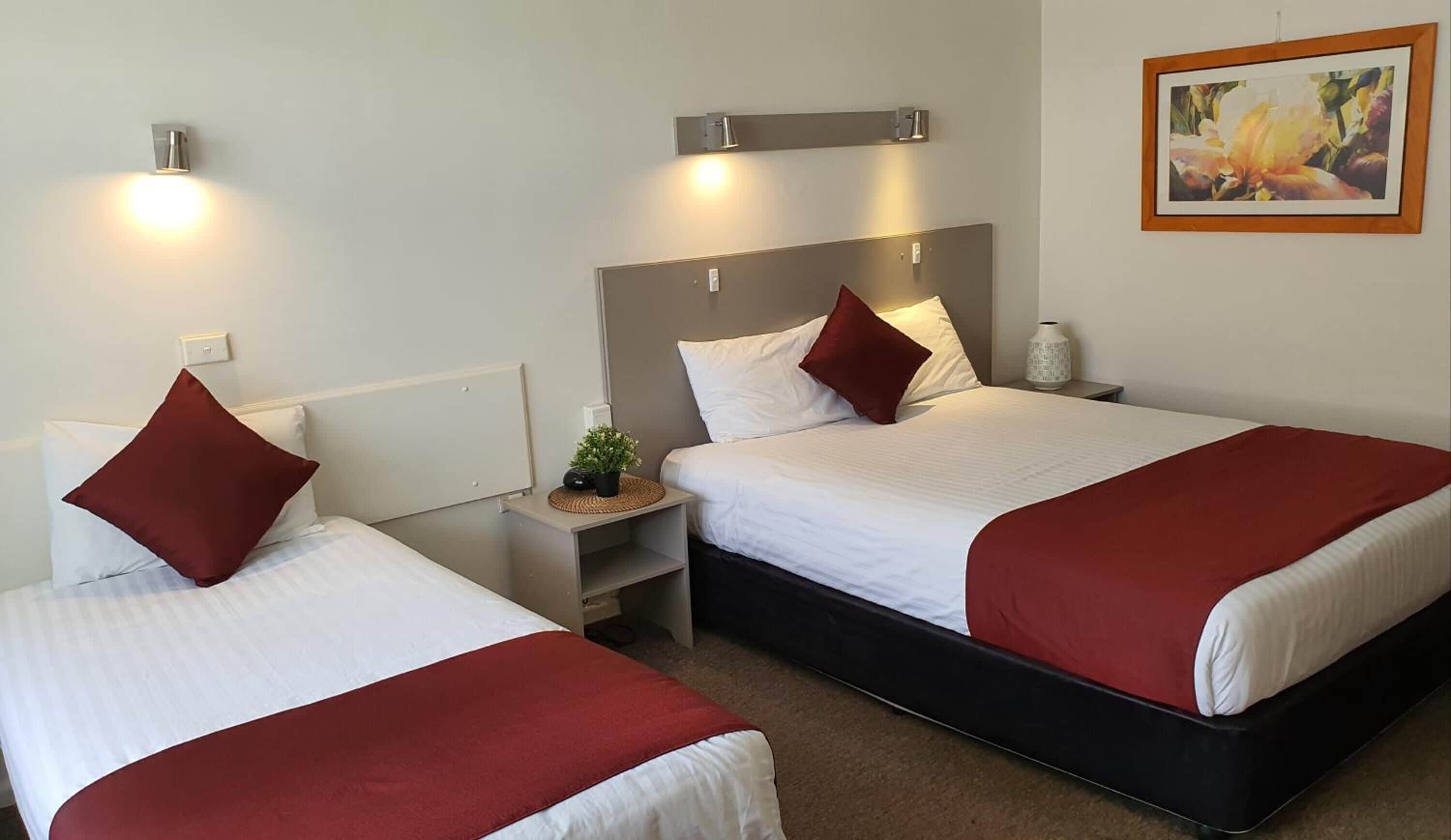 A hotel room with two beds, each with a red and white bedspread, a nightstand with a lamp, and a small plant.