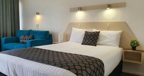 A hotel room with a large bed, a blue armchair, a bedside table with a plant, and wall-mounted lights.