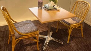 Une pièce avec une fenêtre donnant sur un immeuble et des voitures garées. On y trouve une table avec une boîte de mouchoirs et une petite plante. Deux chaises en osier avec des coussins sont placées autour de la table.