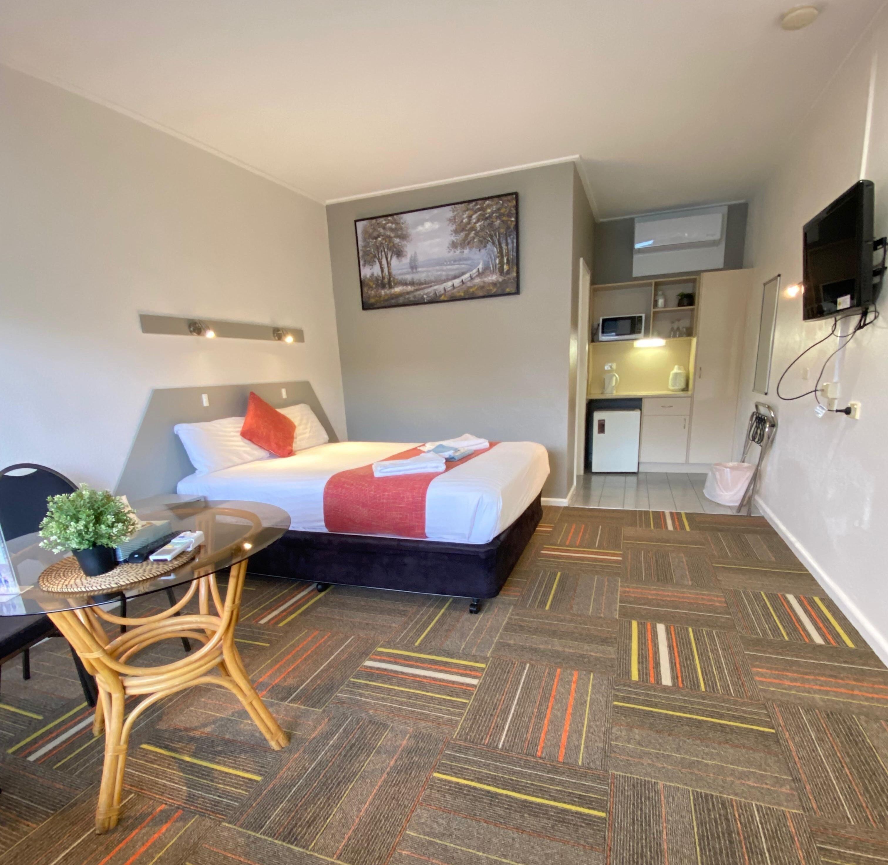 A hotel room with a bed, a small desk with a chair, a potted plant, a microwave, and a television mounted on the wall.