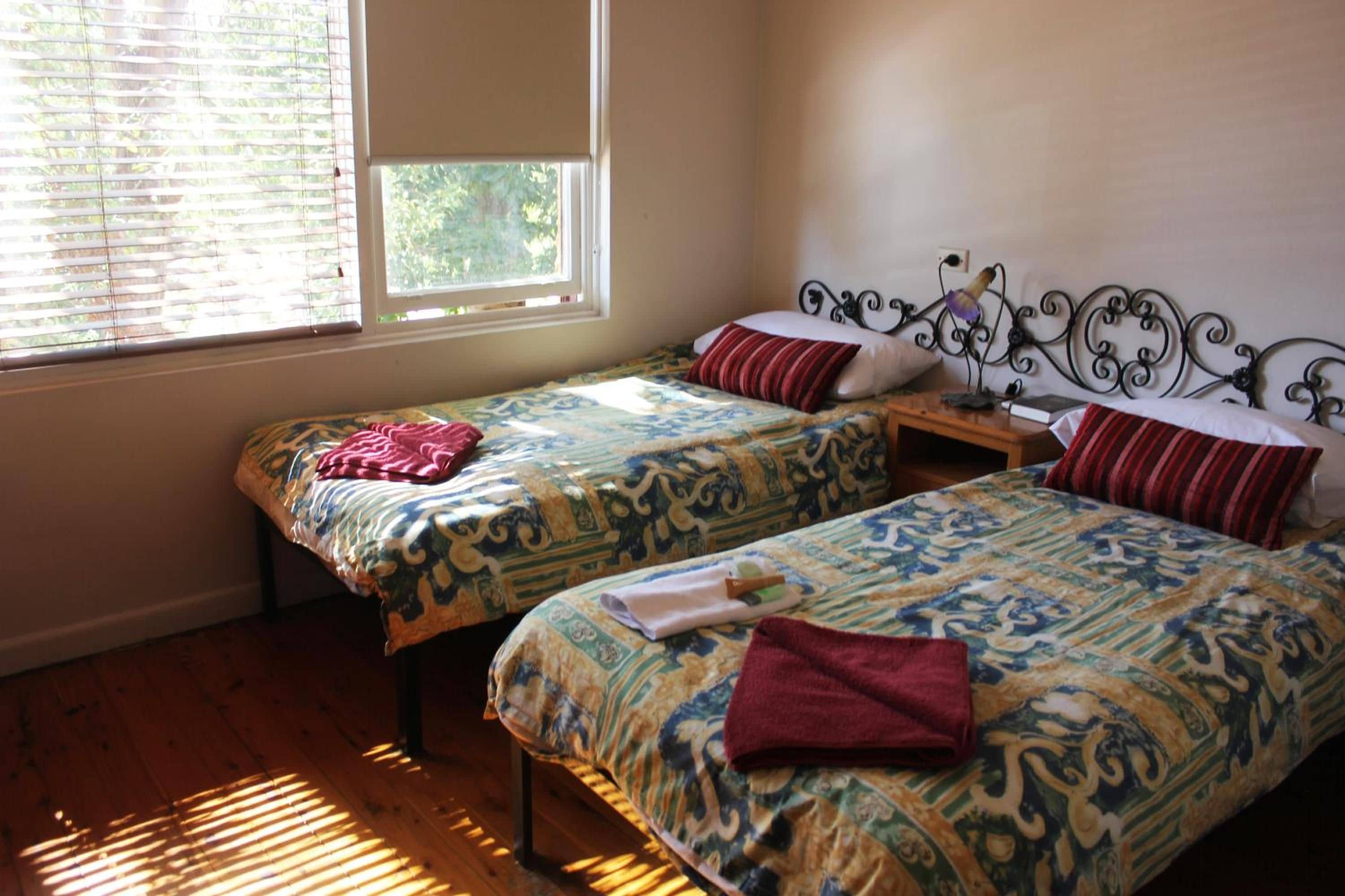 A room with two single beds, each with patterned bedding and pillows, a bedside table with a lamp, and a window with blinds.