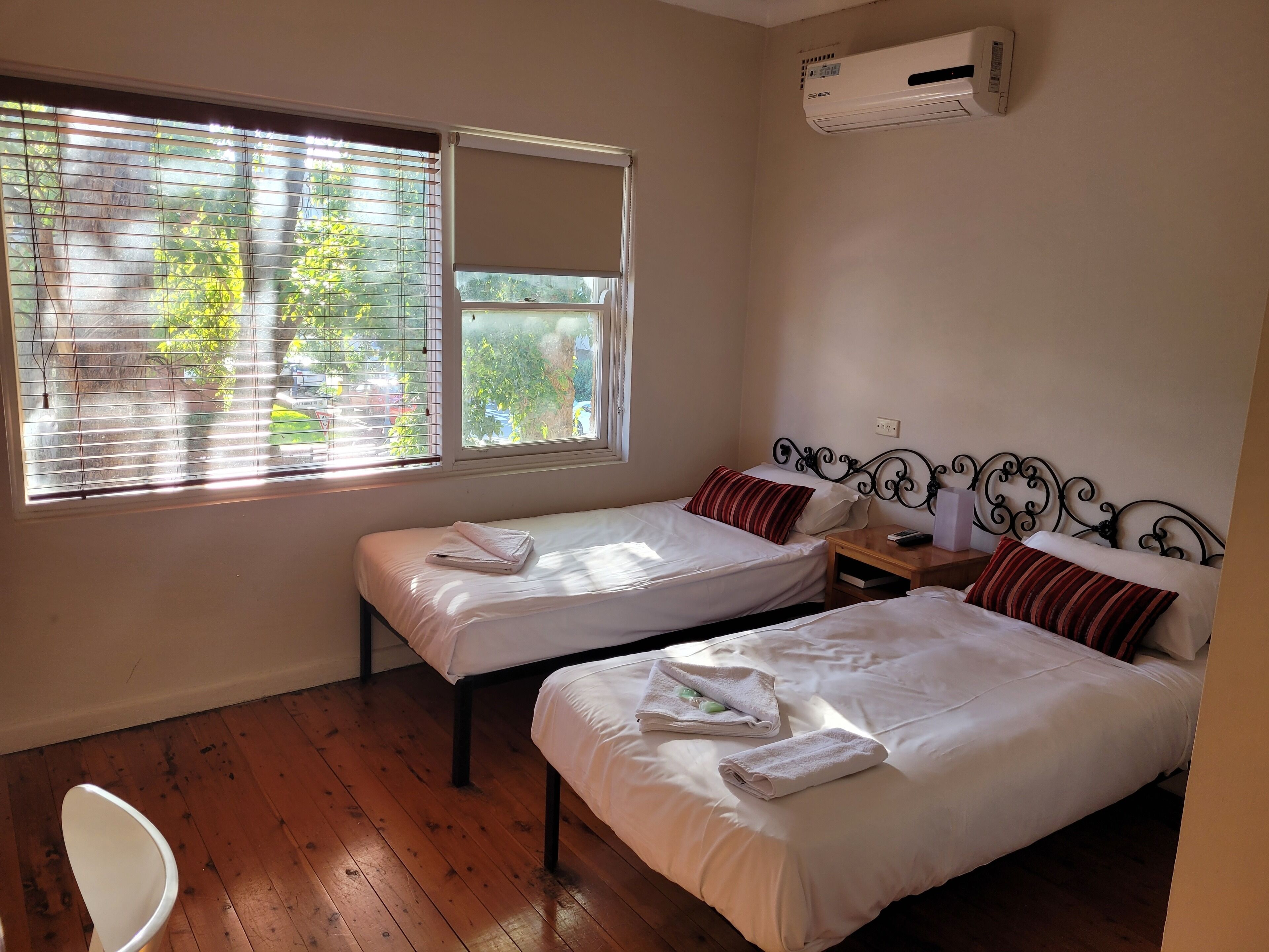 A hotel room with two single beds, a small bedside table with a lamp, a window with blinds, and an air conditioning unit on the wall.