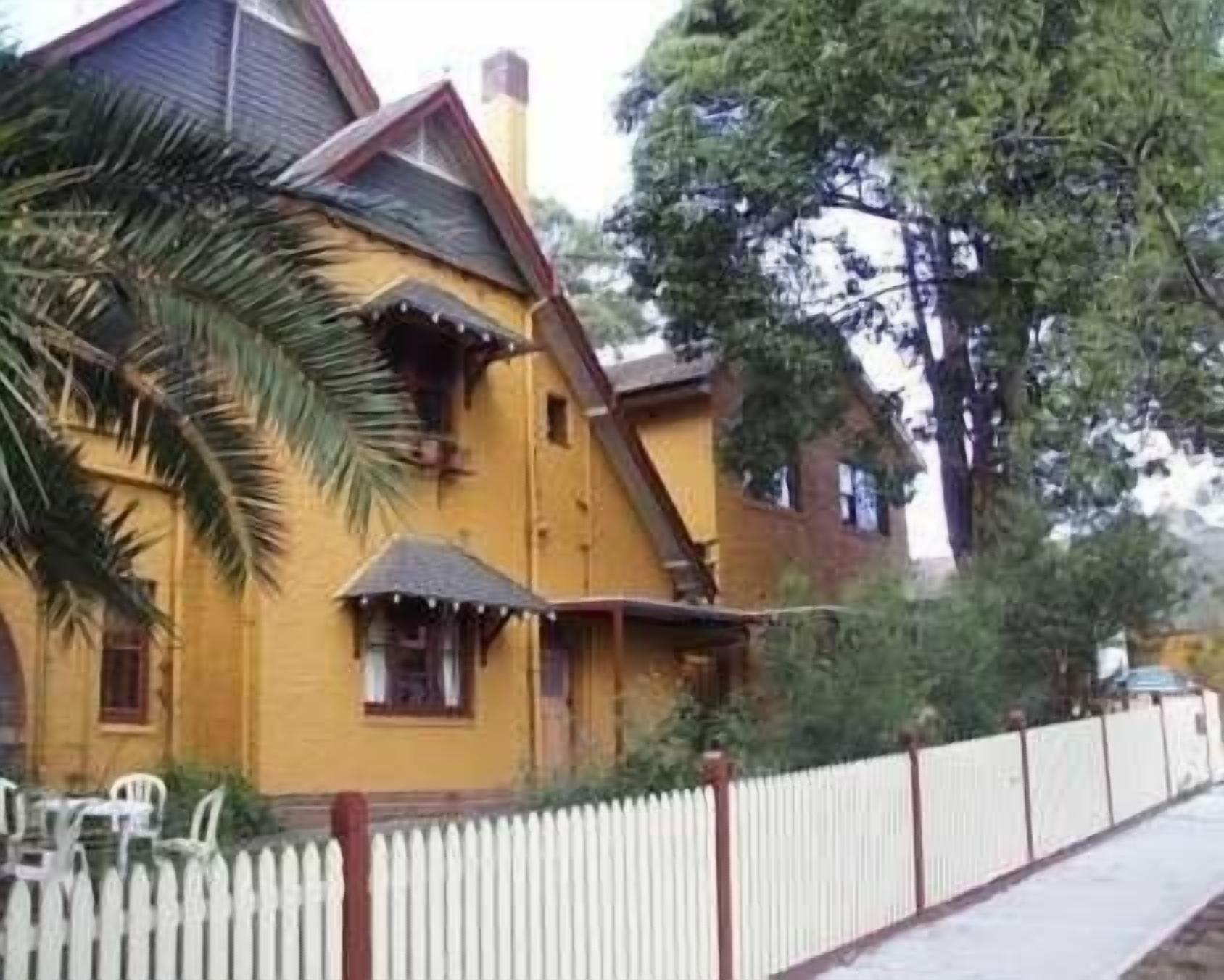 A yellow house with a white picket fence, a porch, and a large tree in the background.