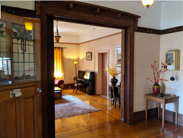 A room with a piano, a sofa, a table with a vase, and a door with a stained glass window. - Burwood Bed And Breakfast (Burwood)