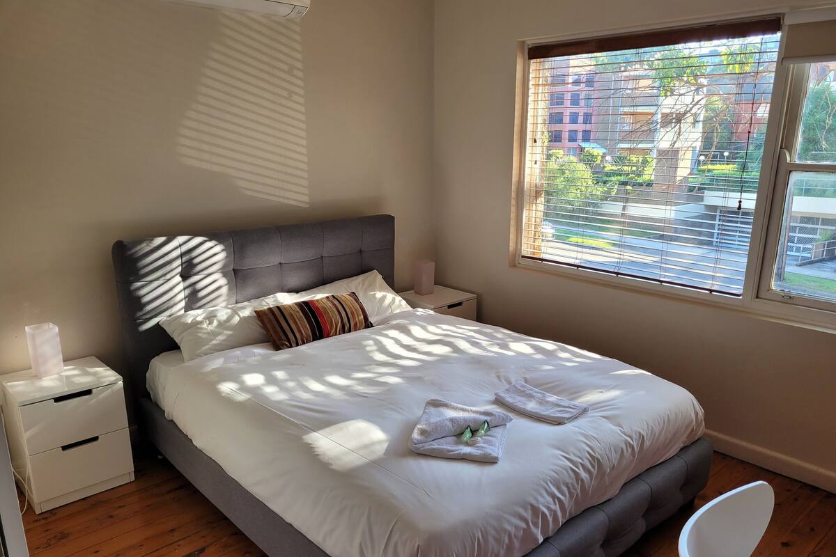 A neatly made bed with white linens and a striped pillow, a bedside table with a lamp, and a window with blinds.