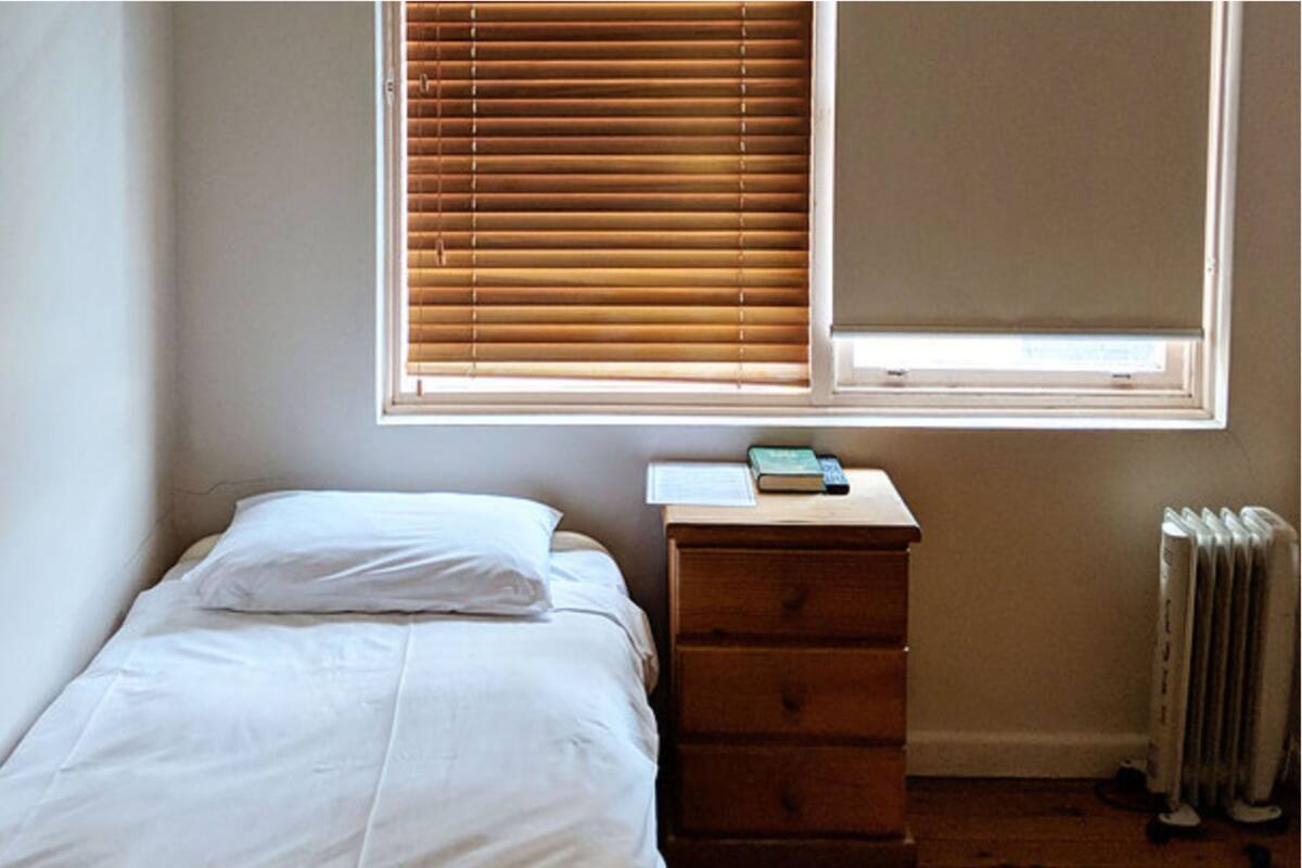 A small bedroom with a single bed, a wooden dresser, a radiator, and a window with blinds.
