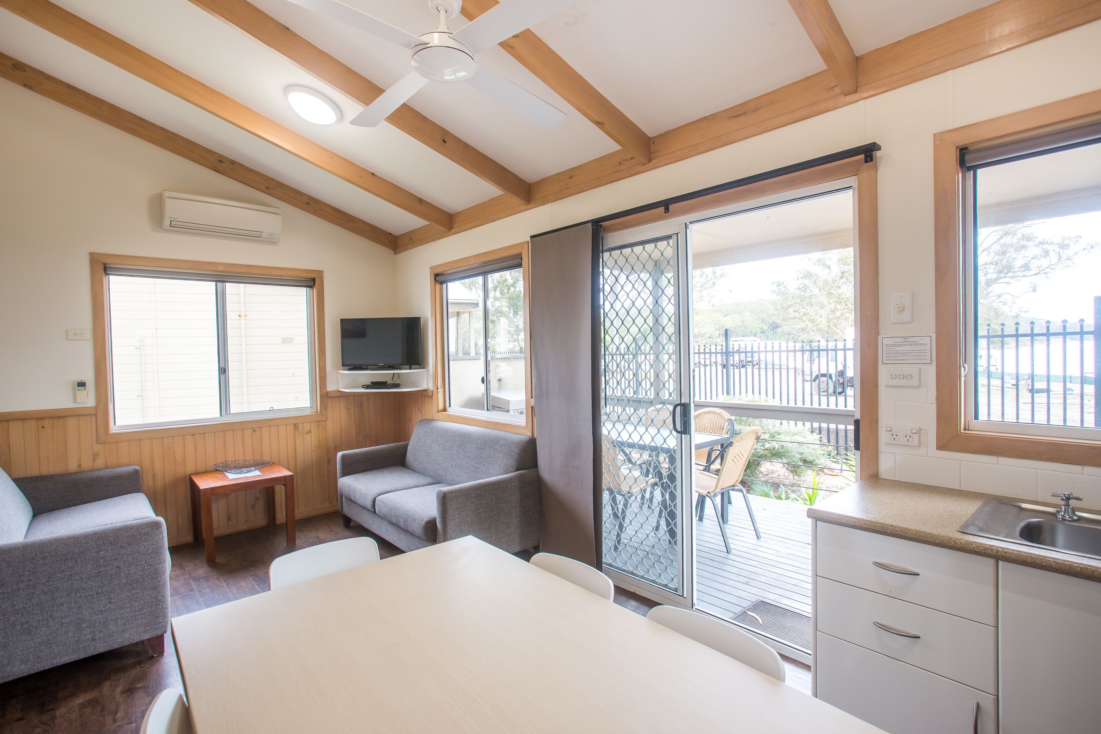 Una habitación con techo de madera, televisor, puerta corrediza de vidrio a un espacio exterior, una pequeña mesa y una kitchenette con lavabo.