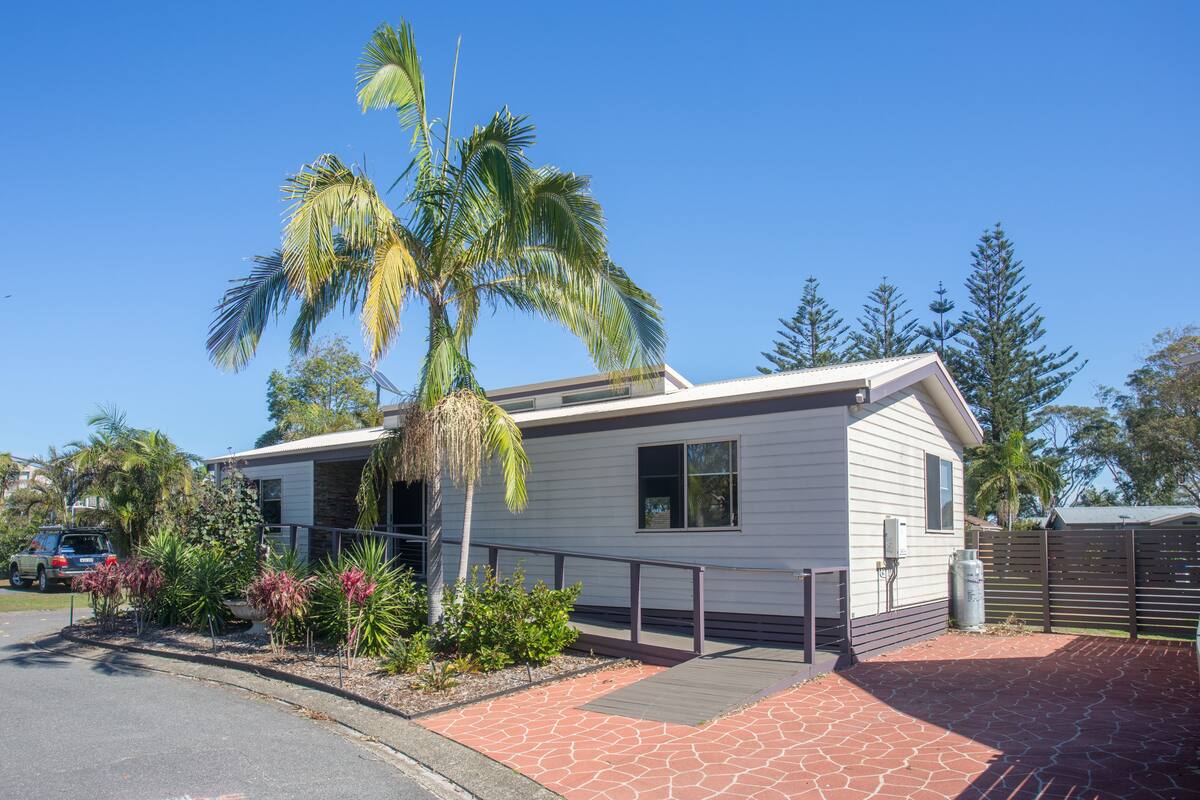A single-story house with a covered porch, surrounded by tropical plants and a paved driveway.