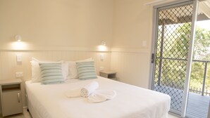 Una cama bien tendida con ropa blanca y almohadas a rayas, una mesita de noche pequeña y una puerta que da a un balcón exterior.