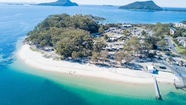 Vista aérea de una zona costera con playa de arena, un conjunto de edificios y dos islas destacadas al fondo.