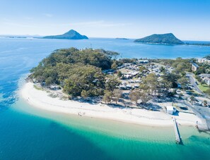 An aerial view of a coastal area with a sandy beach, a cluster of buildings, and two prominent islands in the background. - Halifax Holiday Park (Nelson Bay)