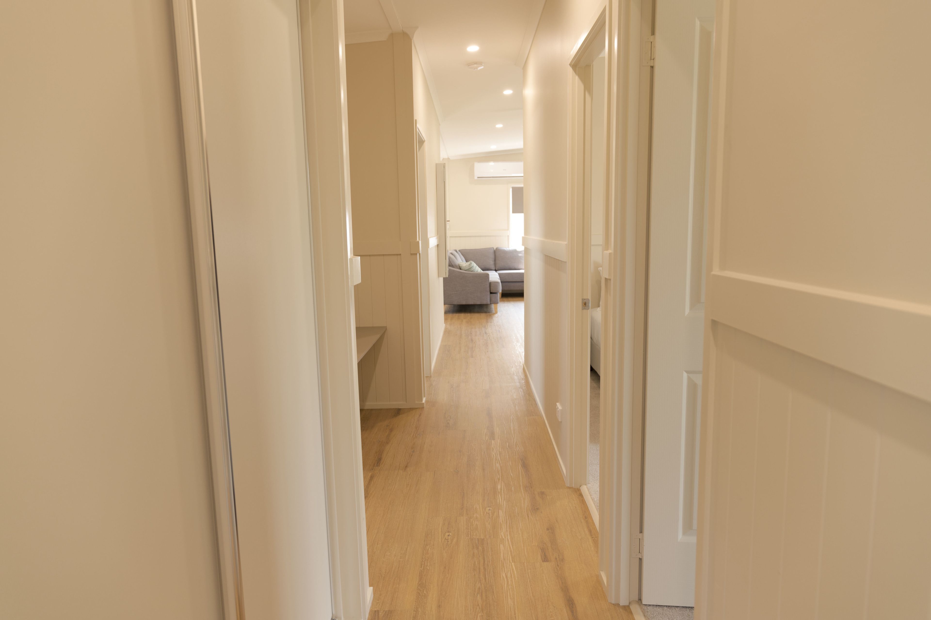 Un couloir étroit, doté d’un plancher en bois et de murs blancs, mène à un espace de vie comprenant un canapé gris et une petite table.