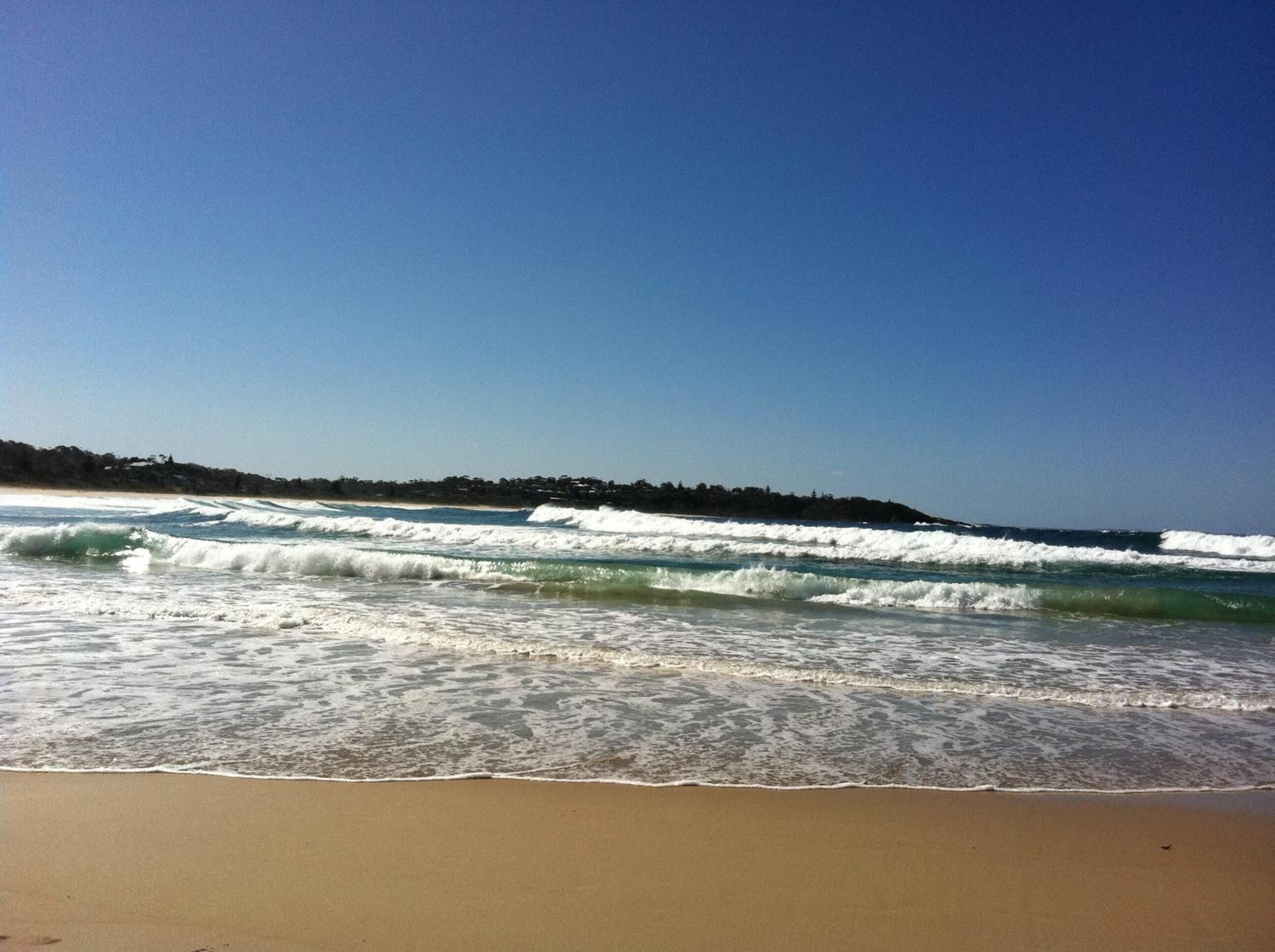 A beach with waves, a clear sky, and a distant shoreline.