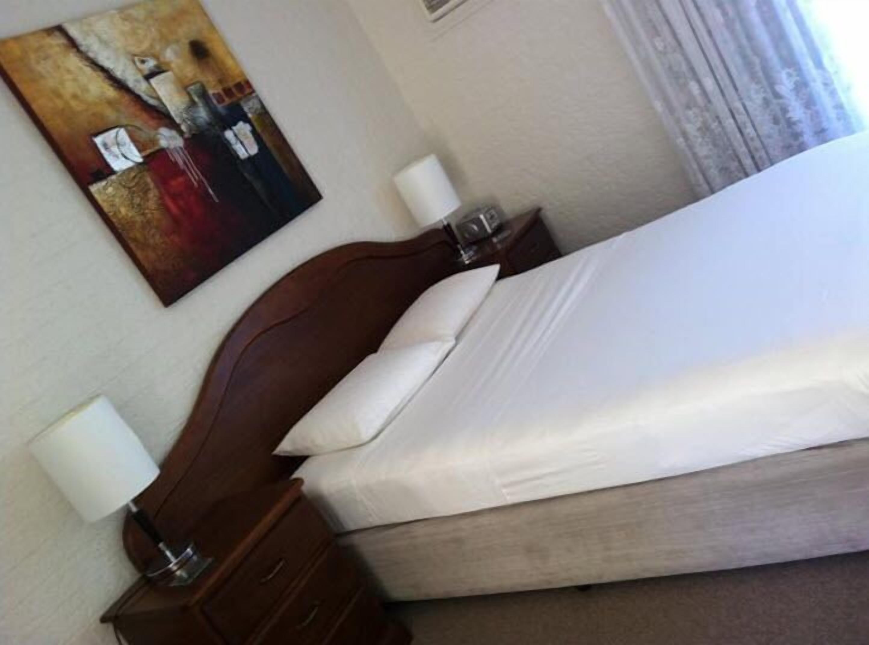 A hotel room with a double bed, a wooden headboard, a nightstand with a lamp, and a painting on the wall.