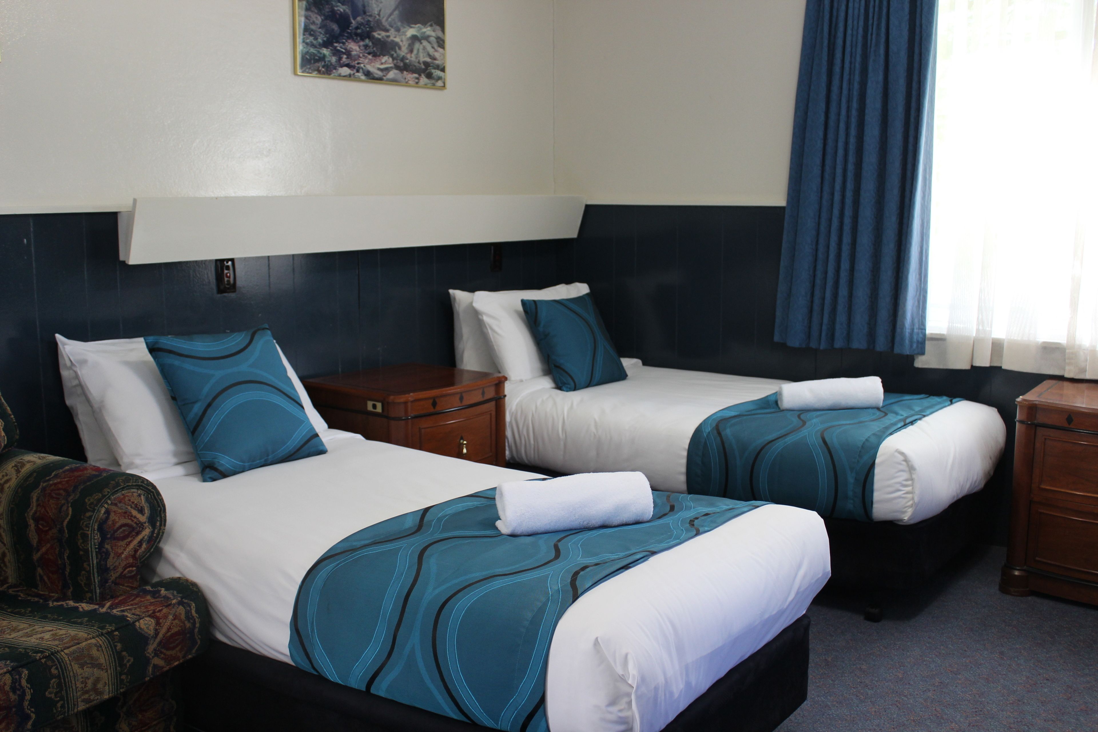 Een hotelkamer met twee bedden, elk met een blauw-witte spreideken, een houten nachtkastje en een stoel.