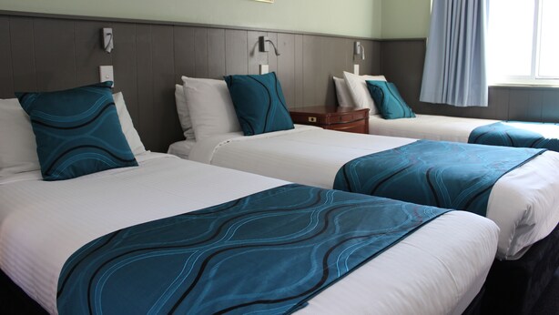 A hotel room with three beds, each with white bedding and teal accents. There is a wooden headboard, a small wooden nightstand, and a wall-mounted light fixture. - High Mountains Motor Inn (Blackheath)
