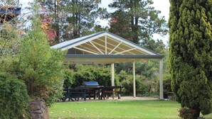A covered outdoor area with a barbecue setup, surrounded by trees and a stone wall. - High Mountains Motor Inn (Blackheath)