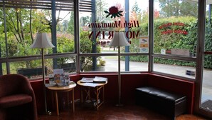A waiting area with a bench, two tables with reading materials, and large windows offering a view of trees and a parking lot. - High Mountains Motor Inn (Blackheath)