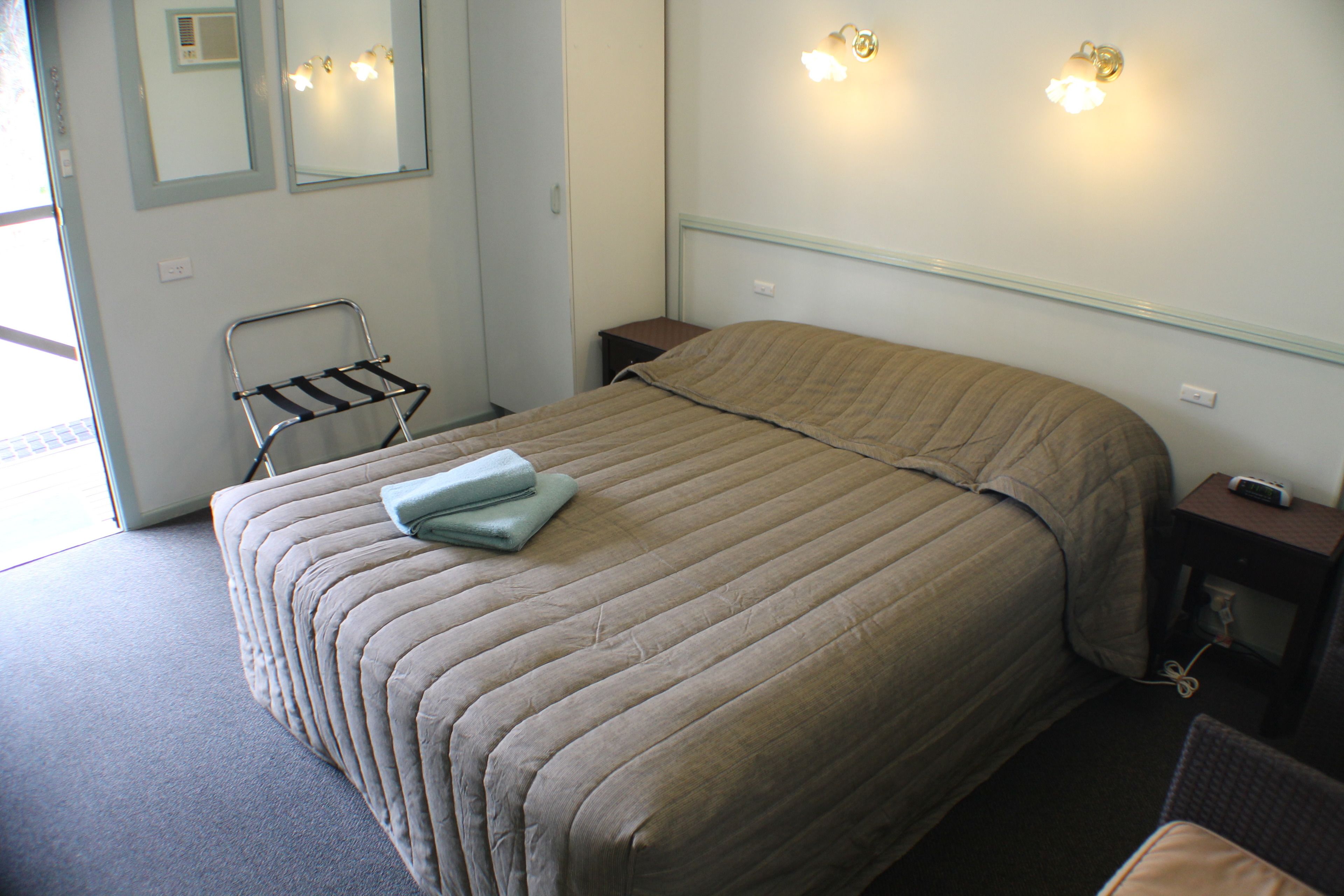En pænt redt seng med et beige dynebetræk, et sengebord med lampe, og et spejl på væggen.