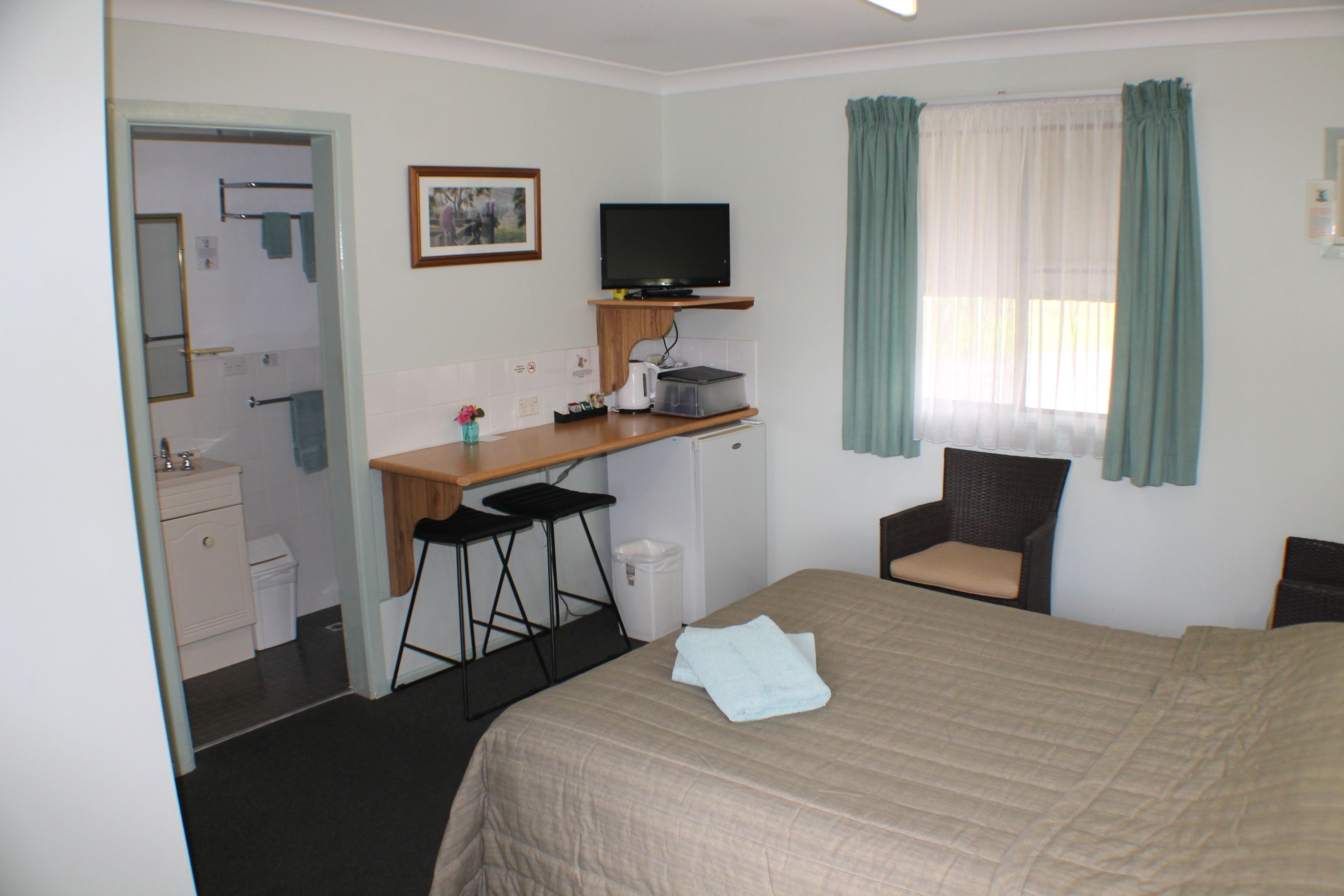 A hotel room with a bed, a desk with a computer, a chair, a small kitchenette, and a bathroom.