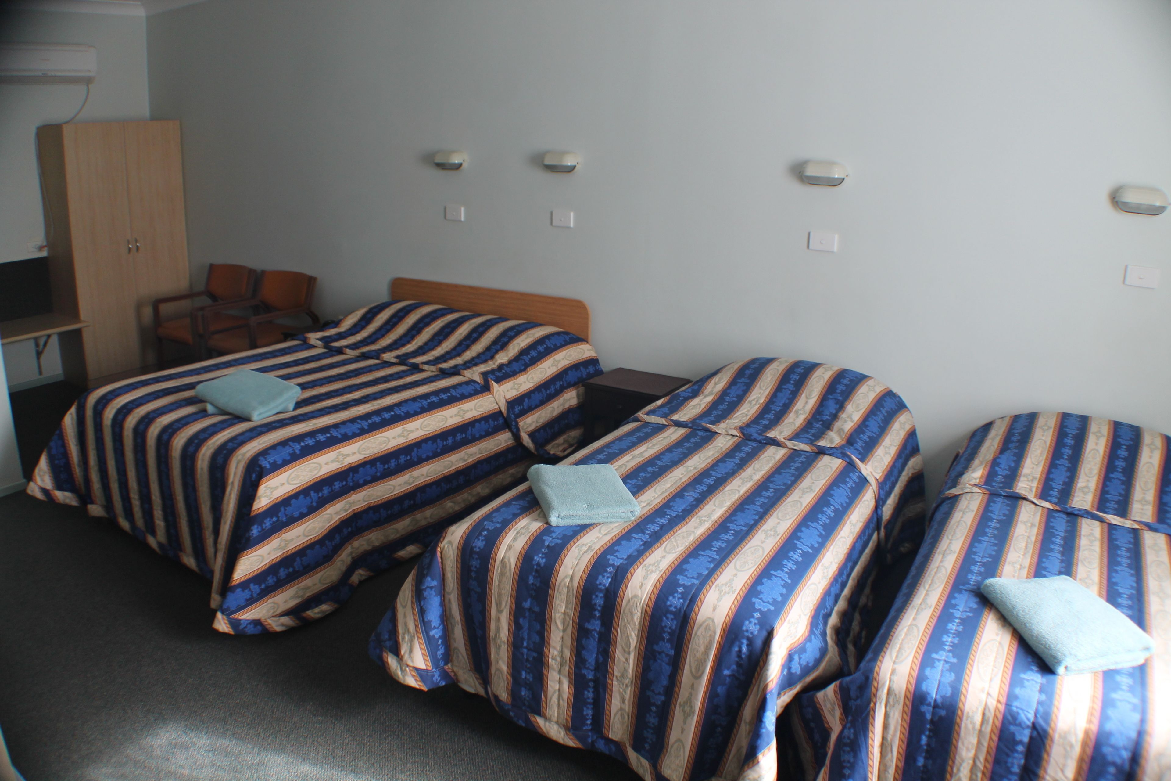Une chambre d’hôtel avec trois lits, chacun doté de draps à rayures et d’un oreiller. On y trouve une armoire en bois et un bureau avec une chaise.