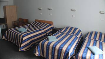 Hotelkamer met drie bedden, elk voorzien van gestreept beddengoed en een kussen. Er is een houten garderobe en een bureau met een stoel.