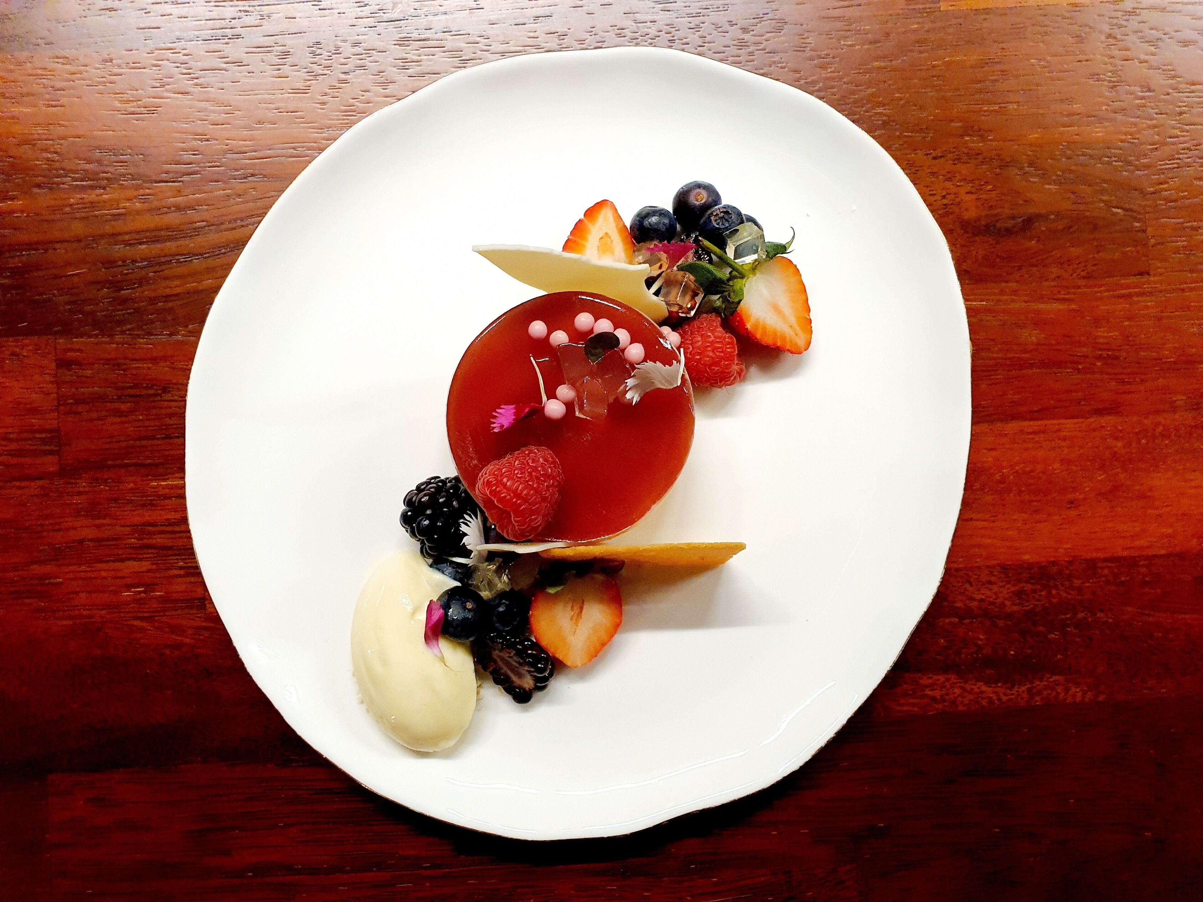 Un plato blanco con un postre de gelatina roja, fresas, frambuesas, zarzamoras y una bola de helado de vainilla.