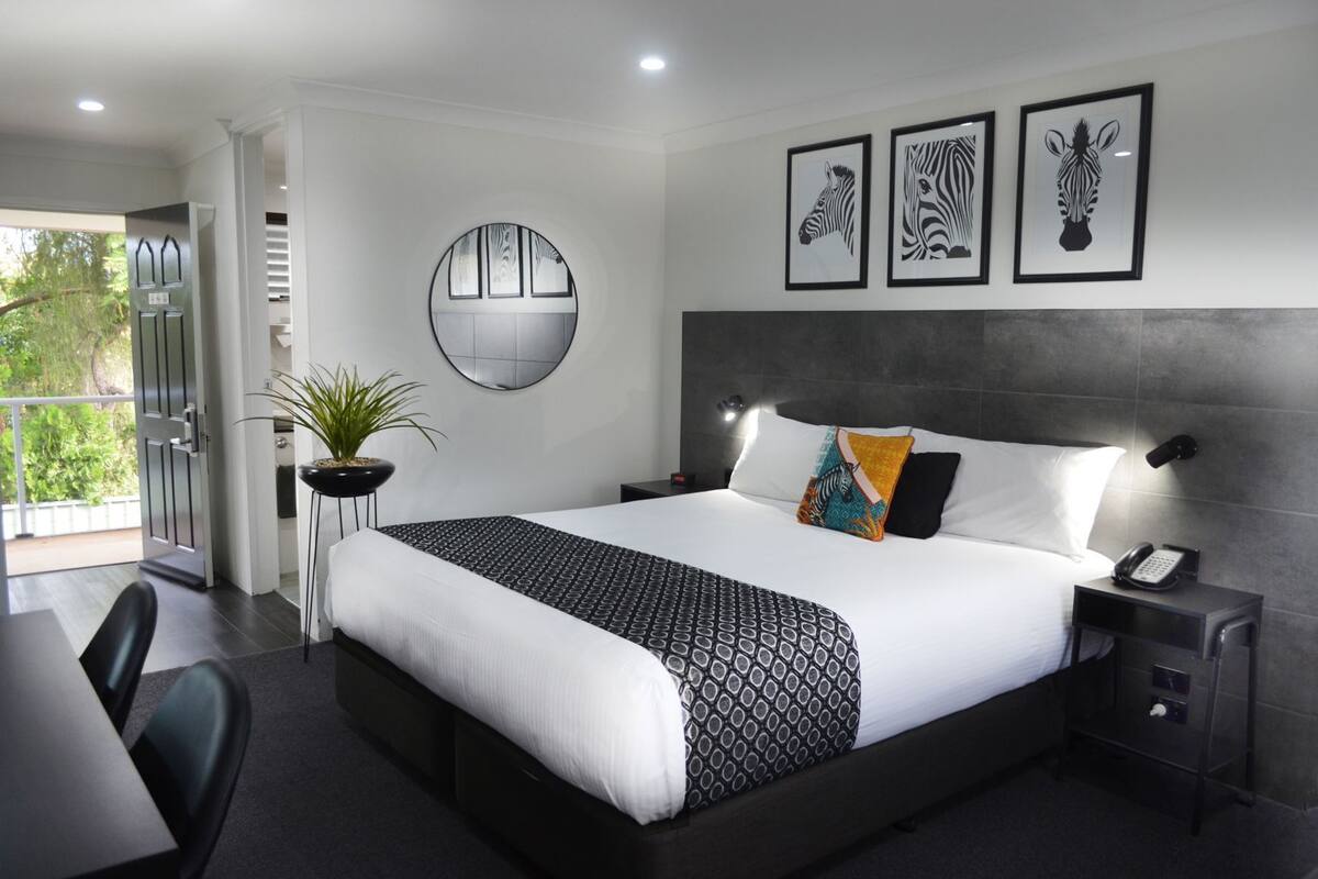 A modern hotel room with a large bed, a desk, a potted plant, and framed artwork on the wall.