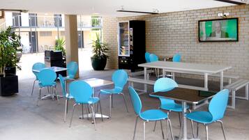 An outdoor dining area with blue chairs and tables, a vending machine, and potted plants.