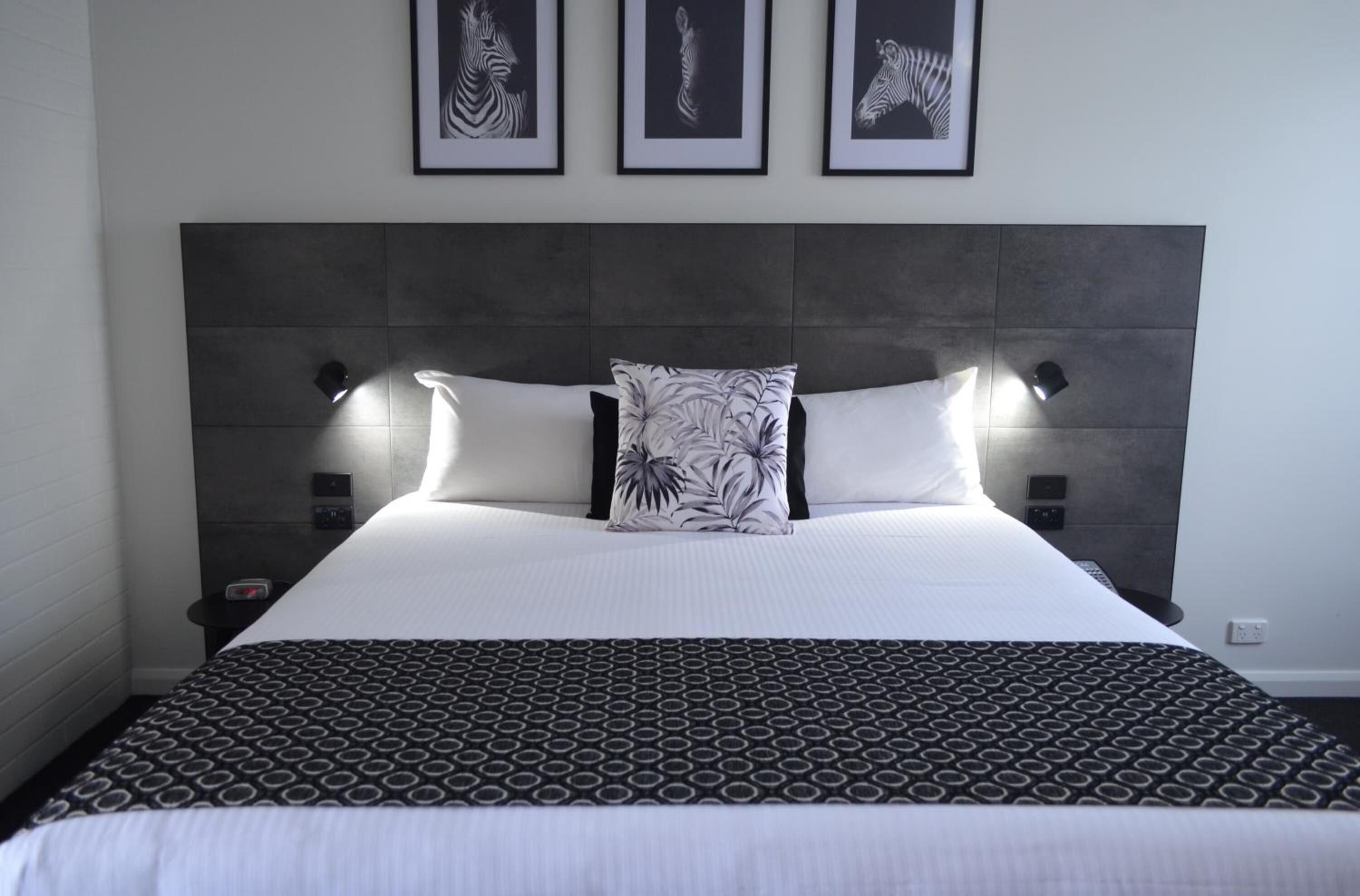 A neatly made bed with a patterned bedspread and two pillows, a headboard with wall-mounted lights, and three framed artworks above the bed.