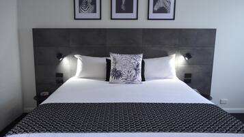 A neatly made bed with a patterned bedspread and two pillows, a headboard with wall-mounted lights, and three framed artworks above the bed.