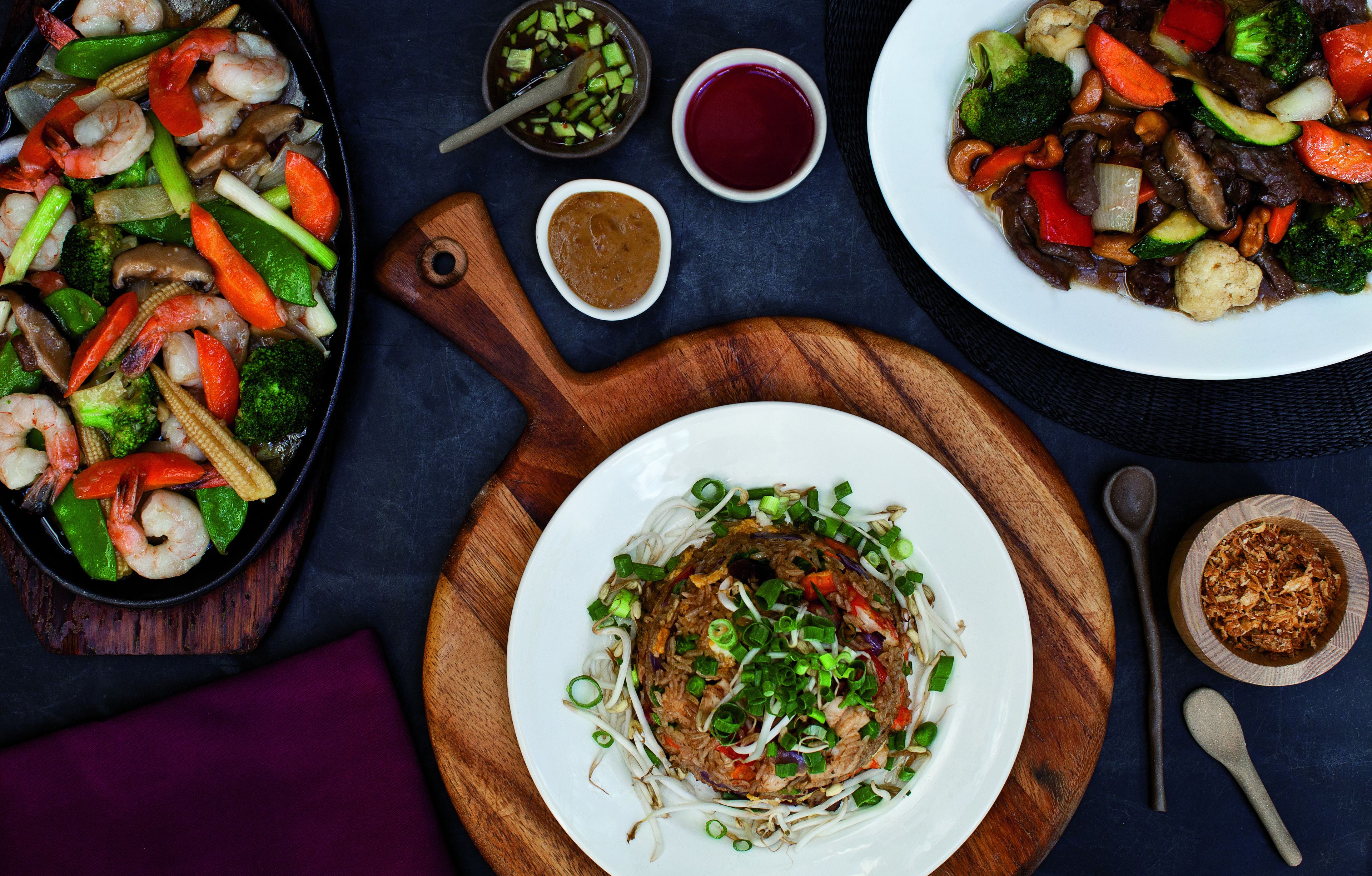 On propose une variété de plats, notamment des légumes et de la viande sautés, servis sur des planches et dans des assiettes en bois.