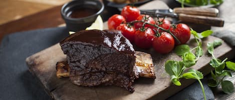 A wooden cutting board with a glazed meat dish, cherry tomatoes, and a bowl of sauce.