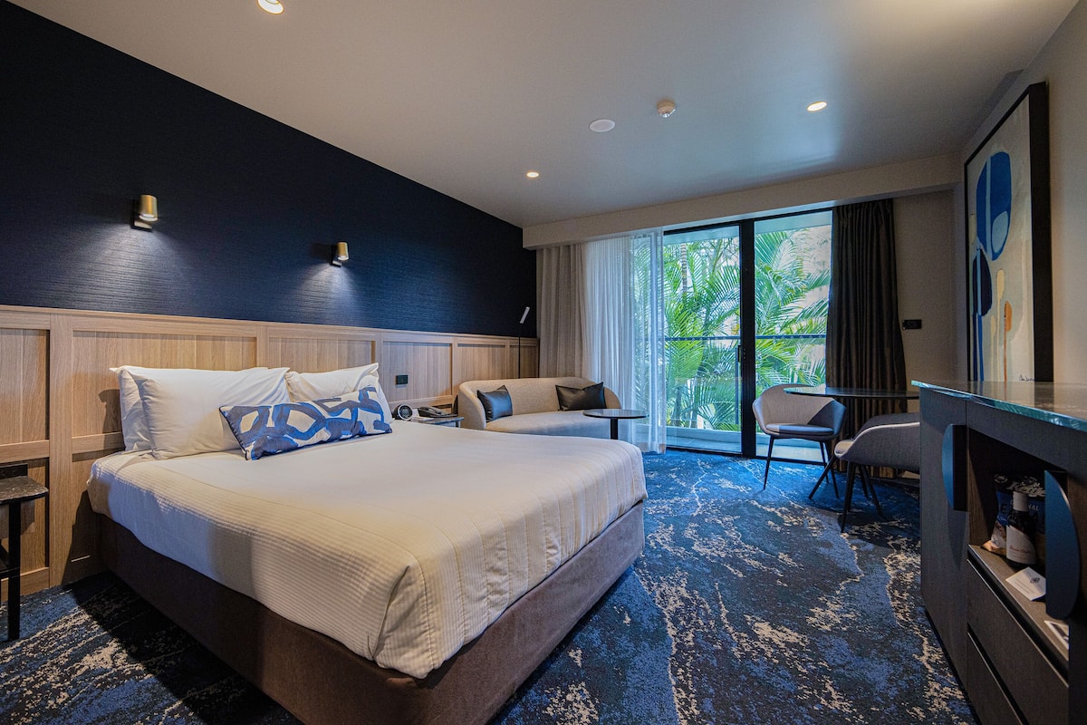 A hotel room with a large bed, two bedside tables, a desk, and a chair. There is a window with curtains and a view of palm trees.