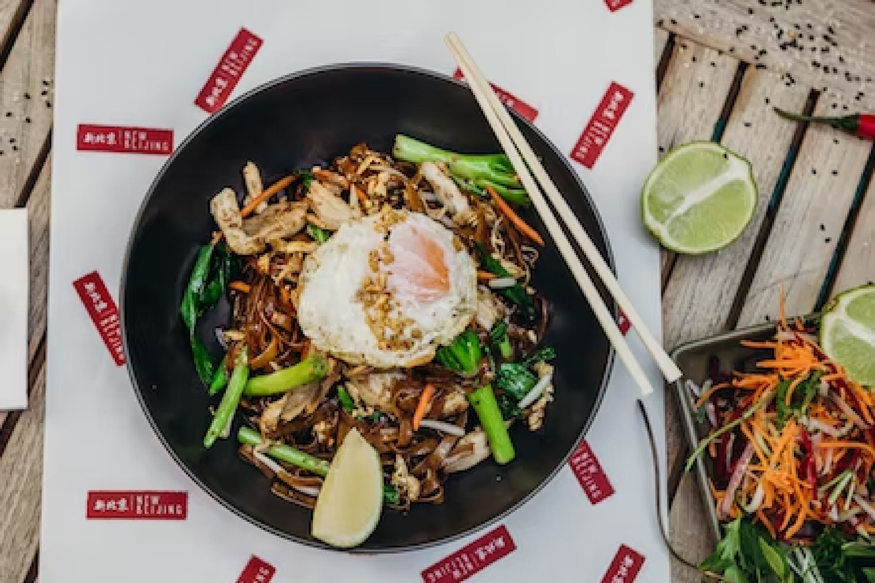 A bowl of stir-fried noodles with vegetables and a fried egg, accompanied by a lime wedge and chopsticks.
