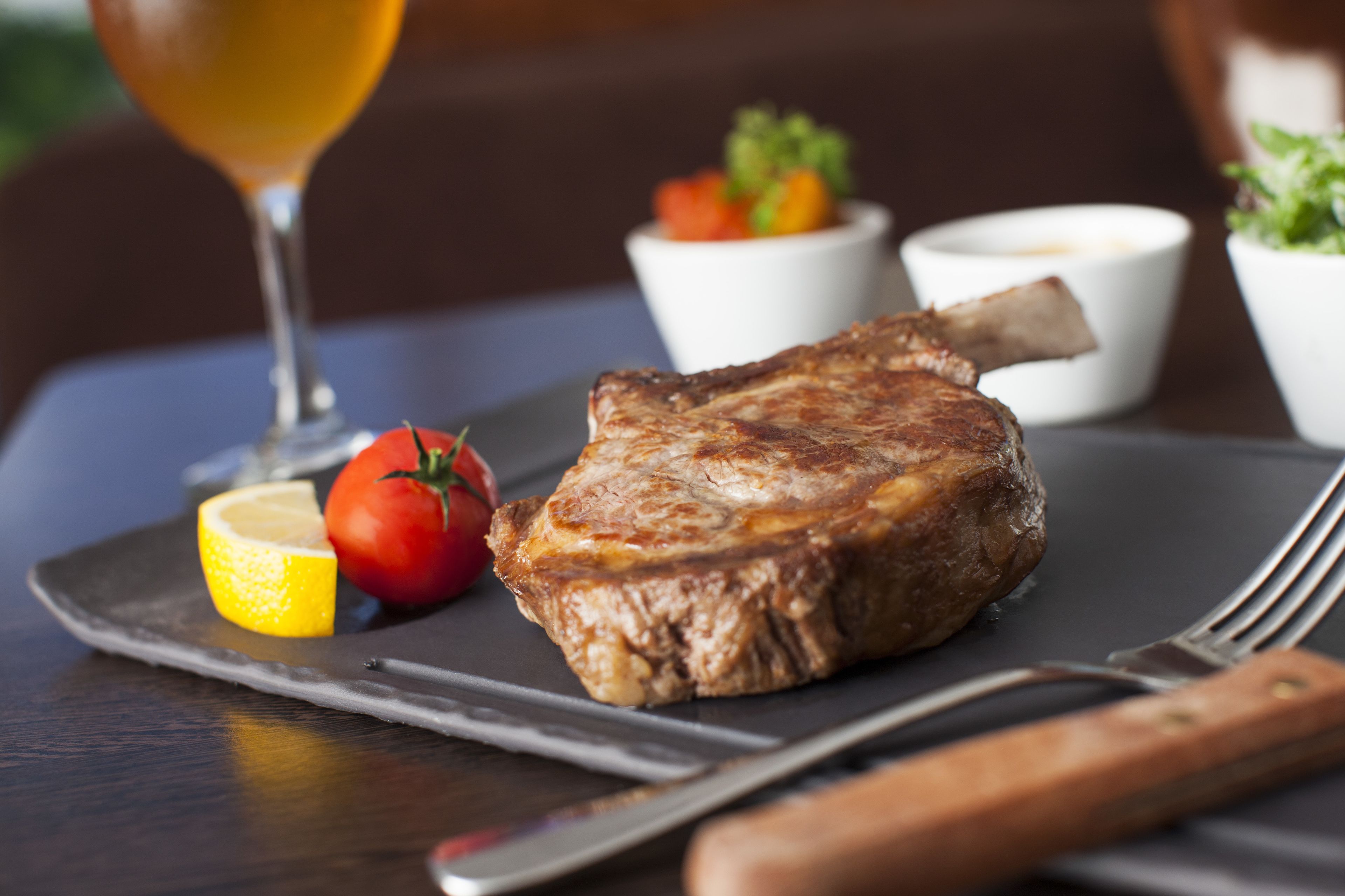 Un steak, une tranche de citron et une tomate sur une assiette, accompagnés d’un verre de bière et d’une fourchette.