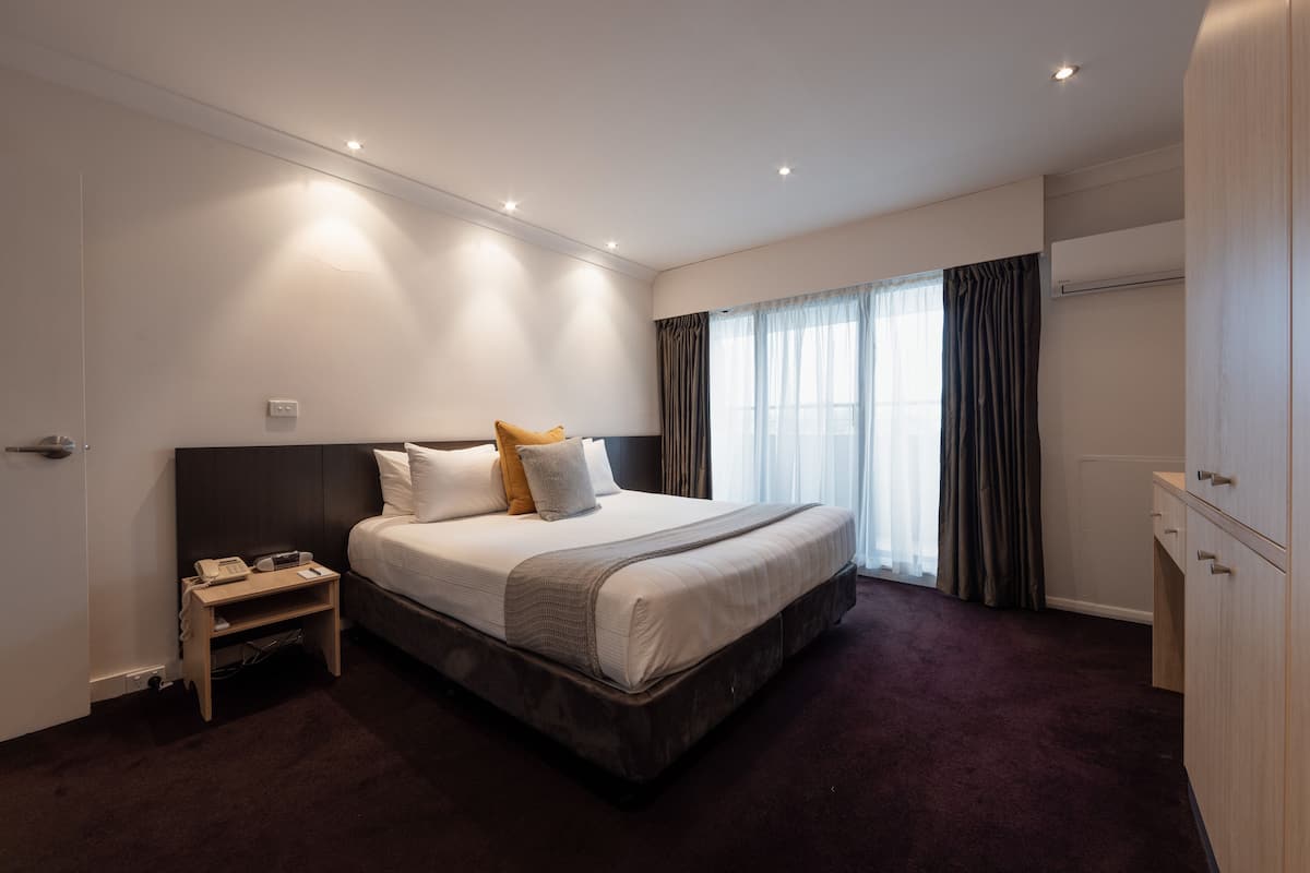 A modern hotel room with a large bed, a bedside table, a wardrobe, and a window with curtains.