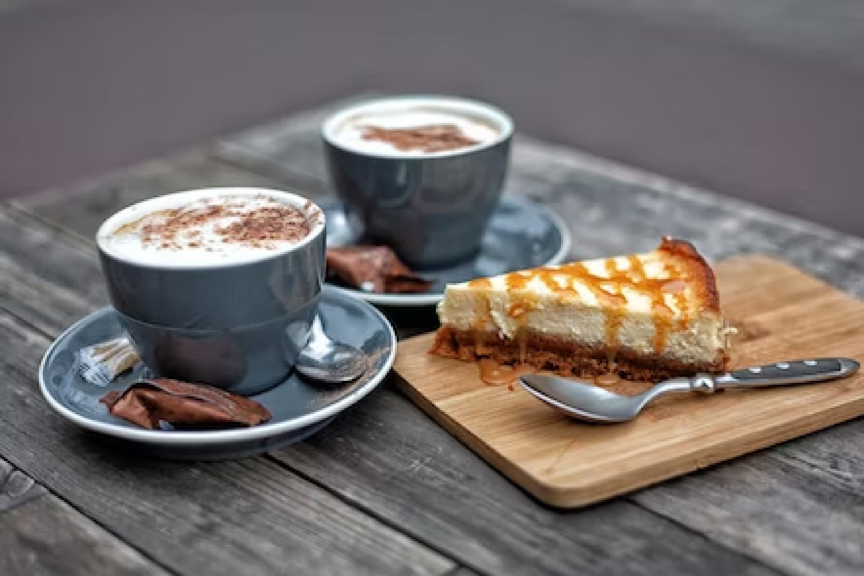 A dessert setup with a slice of cheesecake on a wooden board, two cups of coffee, and a spoon.