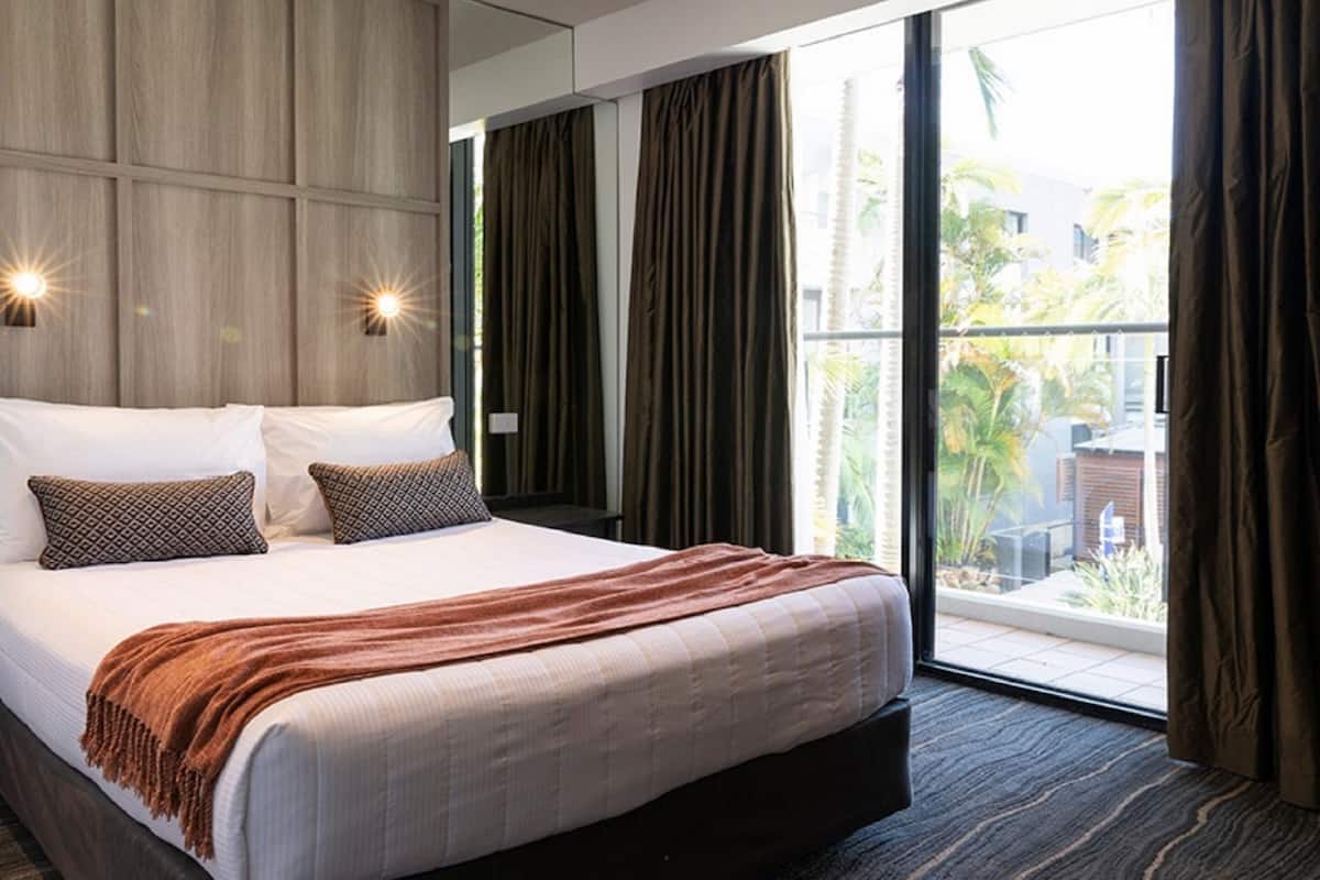 A modern hotel room with a large bed, dark curtains, and a view of a balcony with greenery.