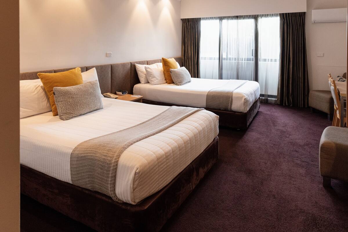 Two double beds with white linens and multiple pillows, a wooden nightstand, a small table, and a chair.