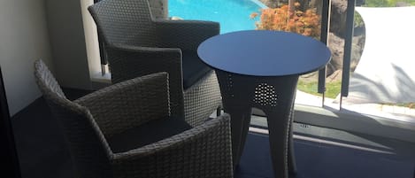 Un balcón con sillas de mimbre y una pequeña mesa, con vistas a una piscina y abundante vegetación.