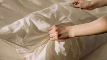 A bed with white sheets and pillows, a person's feet on the bed, and a hand adjusting a pillow.