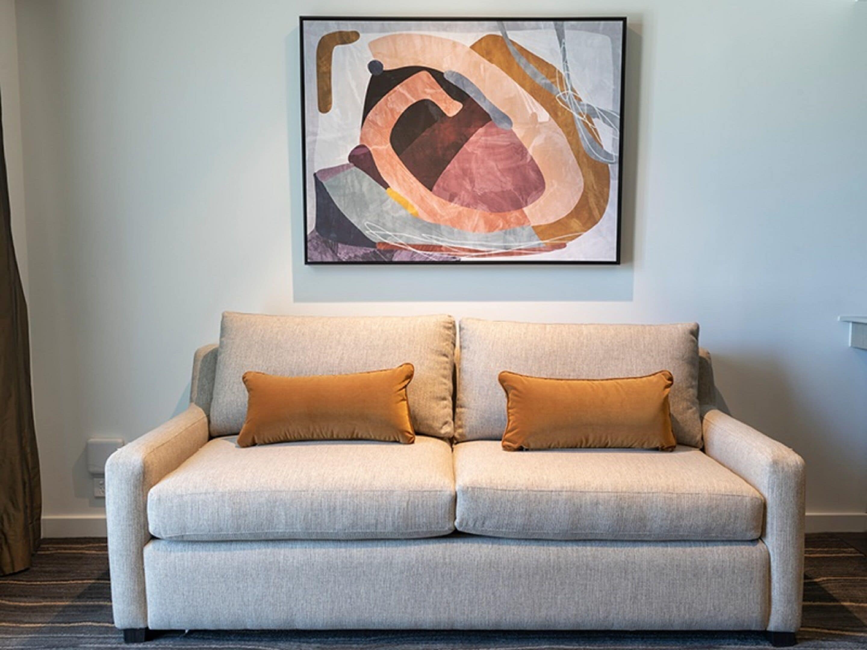 A neutral-colored sofa with two matching cushions, a framed abstract painting above it, and a carpeted floor.
