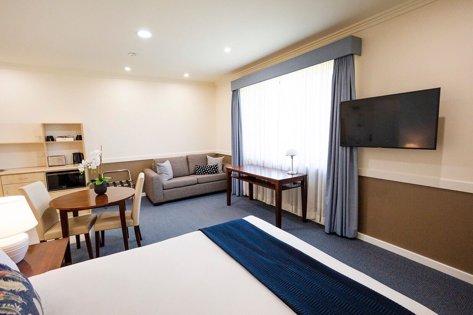 A hotel room with a bed, a sofa, a dining table, a TV, and a kitchenette.