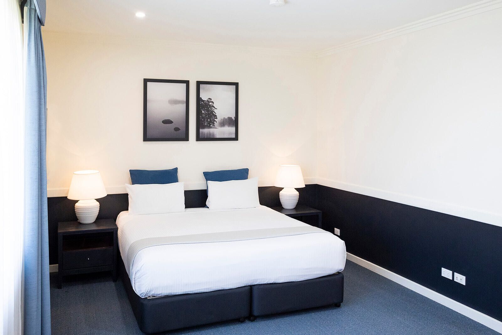 A modern hotel room with a large bed, two bedside tables, and two wall-mounted picture frames.