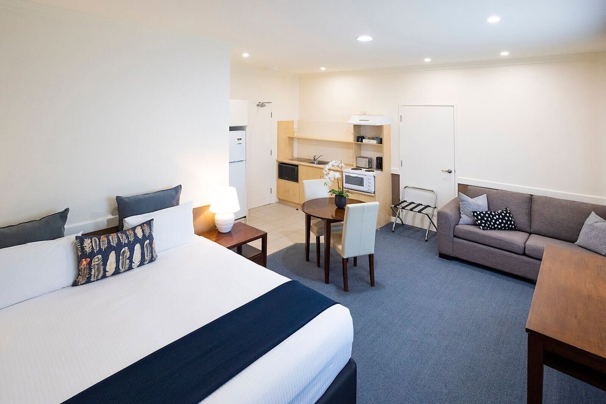 A hotel room with a bed, a desk, a chair, a sofa, and a kitchenette.