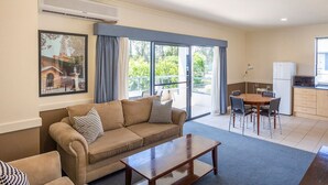 Sala de estar con sofá, mesa de centro y comedor con sillas y una mesa. Hay una zona de cocina con refrigerador y microondas. Se ve un ventanal con cortinas, y un equipo de aire acondicionado está montado en la pared.
