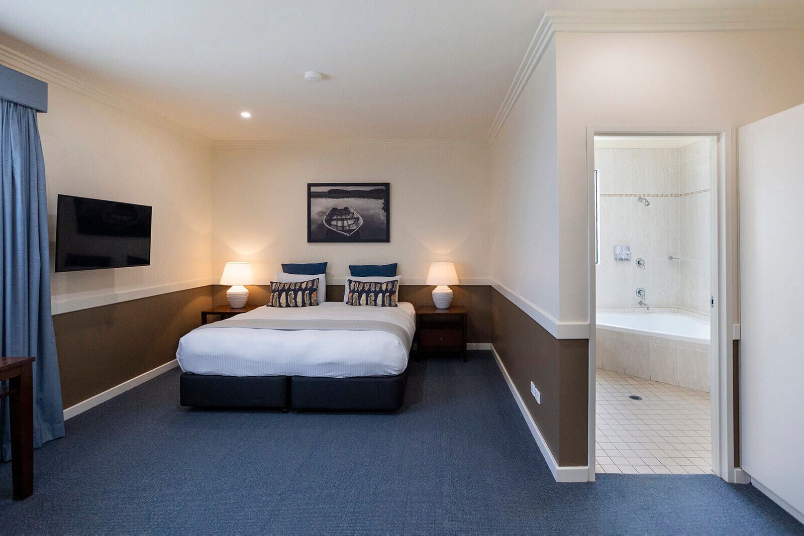 A hotel room with a bed, two bedside tables with lamps, a flat-screen TV, a framed artwork, and a bathroom with a bathtub and shower.