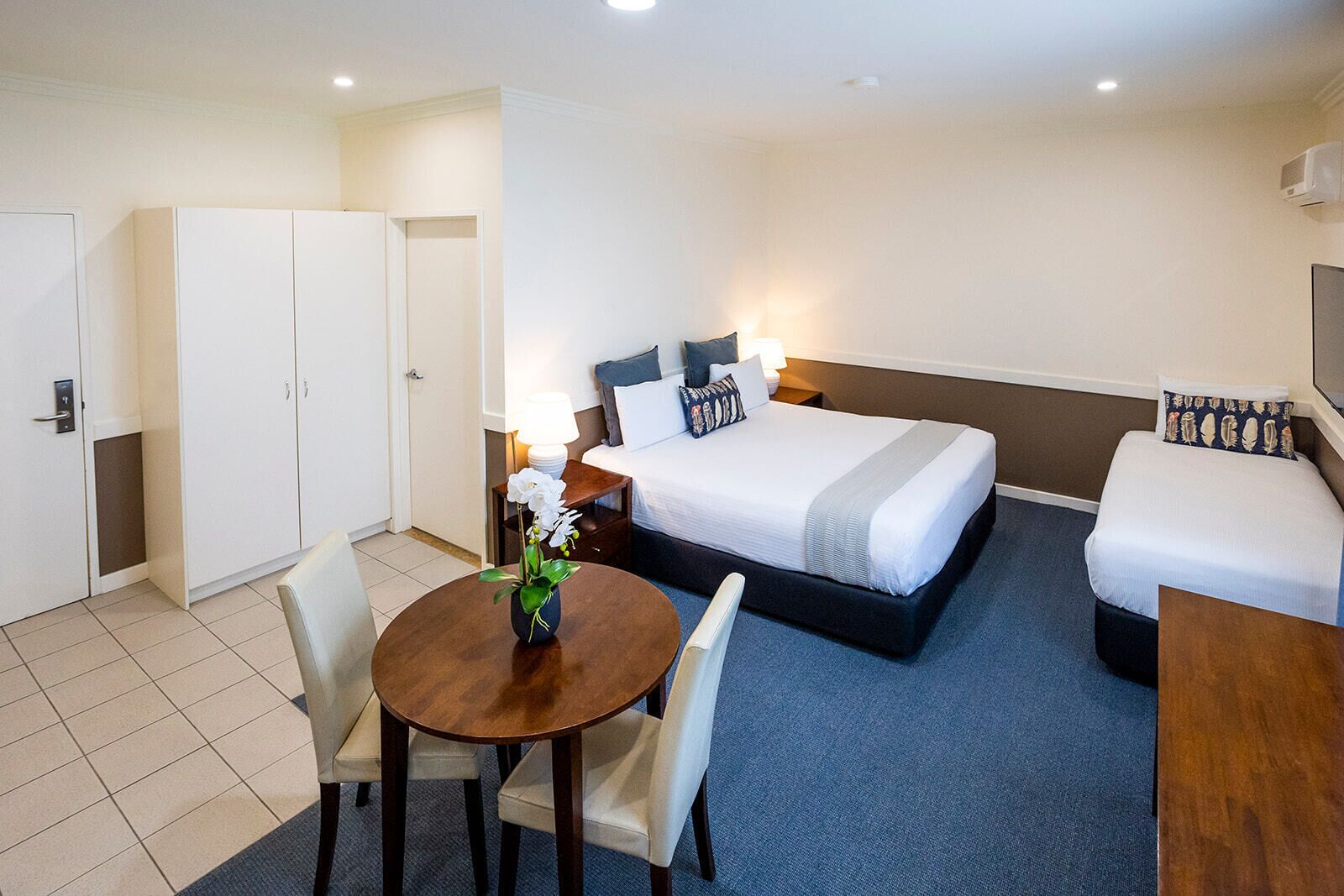 A hotel room with a bed, a desk, a chair, a small table with a vase, and a wardrobe.
